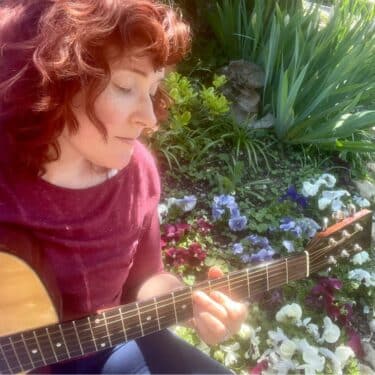A woman with red hair sits in a patch of flowers, playing guitar whilst looking away from the camera