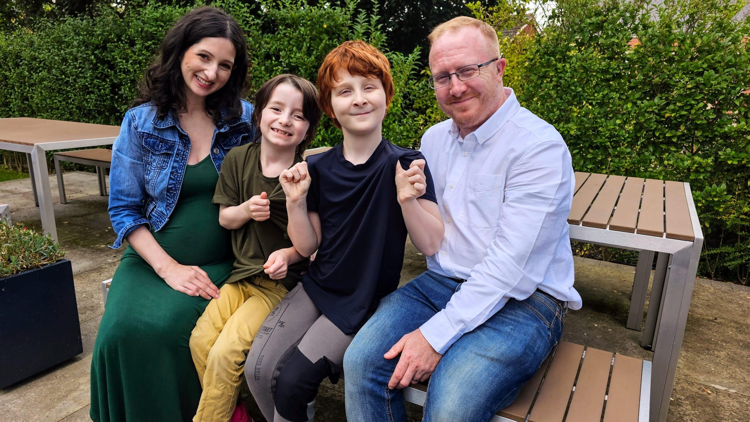 A family of 2 parents and 2 young children smile at the camera. the children are wearing sensory friendly Fidget-T clothing, and the parents are dressed smartly